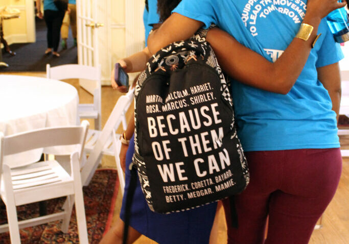 Two students embracing from the back, one wearing a Summer Policy Institute t-shirt, the other with a backpack that says "Because of them we can"